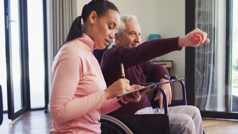 Video-of-happy-biracial-female-physiotherapist-exercising-with-caucasian-senior-man,-making-notes
