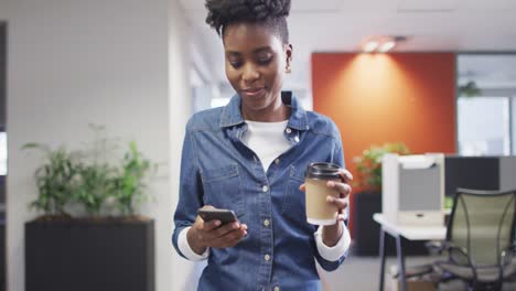Video-Einer-Afroamerikanischen-Geschäftsfrau,-Die-Im-Büro-Spaziert,-Kaffee-Trinkt-Und-Ihr-Smartphone-Benutzt