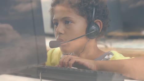 African-american-boy-wearing-phone-headset-using-computer-at-school-against-clouds-in-blue-sky
