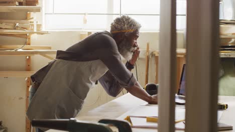 Afroamerikanischer-Zimmermann,-Der-In-Einer-Tischlerei-Mit-Dem-Smartphone-Spricht-Und-Einen-Laptop-Benutzt