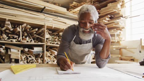 African-american-male-carpenter-talking-on-smartphone-and-taking-notes-in-a-carpentry-shop