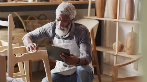African-american-male-carpenter-touching-wooden-chair-frame-and-writing-on-clipboard