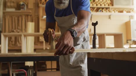 African-american-male-carpenter-cutting-a-wooden-plank-using-a-hand-saw-in-a-carpentry-shop