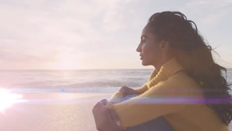 Animation-of-light-spots-over-biracial-woman-sitting-at-beach