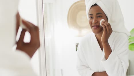 Video-Einer-Glücklichen-Afroamerikanischen-Frau-In-Robe,-Die-In-Den-Spiegel-Schaut-Und-Ihr-Gesicht-Reinigt