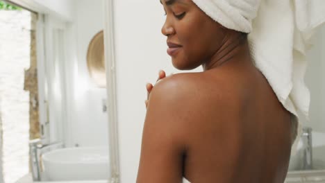 Video-Einer-Glücklichen-Afroamerikanischen-Frau-In-Robe,-Die-Ihre-Haut-Mit-Feuchtigkeit-Versorgt