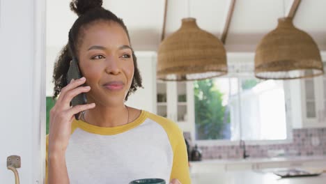 Mujer-Afroamericana-Sosteniendo-Una-Taza-De-Café-Hablando-Por-Teléfono-Inteligente-En-Casa