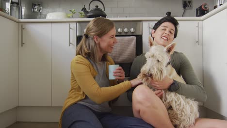 Caucasian-lesbian-couple-holding-coffee-cups-playing-with-their-dog-sitting-in-the-kitchen-at-home