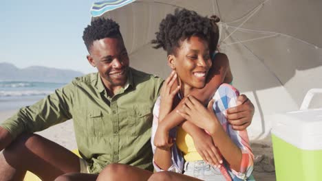 Afroamerikanische-Familie-Lächelt,-Während-Sie-Unter-Einem-Sonnenschirm-Am-Strand-Sitzt