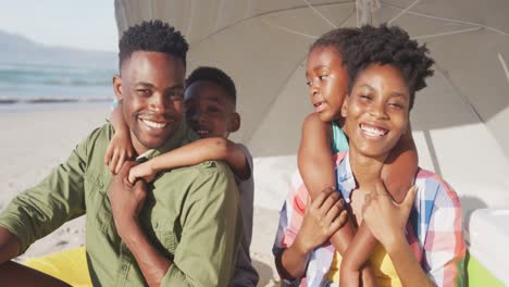 Porträt-Einer-Afroamerikanischen-Familie,-Die-Lächelt,-Während-Sie-Unter-Einem-Sonnenschirm-Am-Strand-Sitzt
