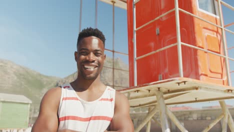 Portrait-of-happy-african-american-male-lifeguard-next-to-his-tower-on-sunny-beach