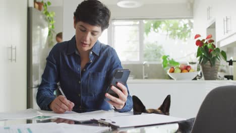 Caucasian-woman-taking-notes-and-calculating-finances-using-smartphone-at-home