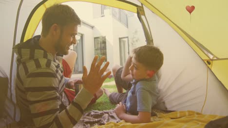 Animation-of-hearts-over-caucasian-father-and-son-high-fiving-in-tent-in-garden