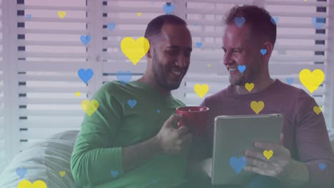 Animation-of-hearts-over-diverse-gay-couple-smiling-and-using-tablet