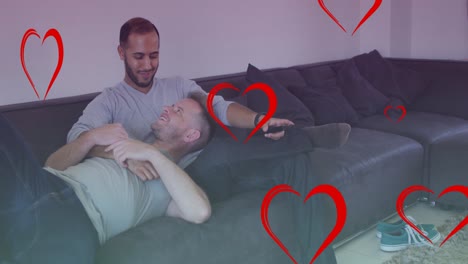 Animation-of-hearts-over-diverse-male-couple-watching-tv