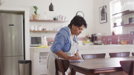 Senior-biracial-woman-wearing-apron-and-cleaning-table-in-kitchen-alone