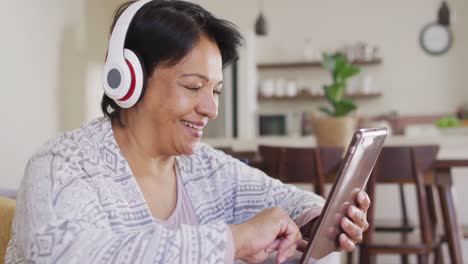 Mujer-Mayor-Afroamericana-Usando-Audífonos-Usando-Una-Tableta-Digital-Sentada-En-El-Sofá-En-Casa