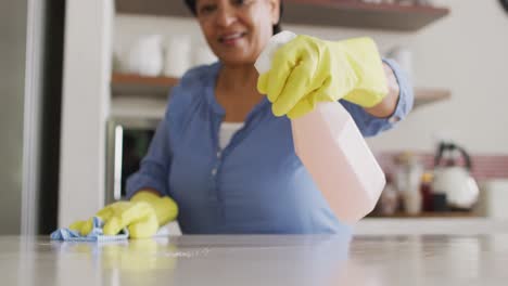 Smiling-senior-biracial-woman-wearing-gloves-and-cleaning-table-in-kitchen-alone