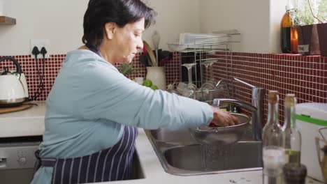 Mujer-Birracial-Mayor-Lavando-Verduras-Y-Usando-Delantal-Solo-En-La-Cocina