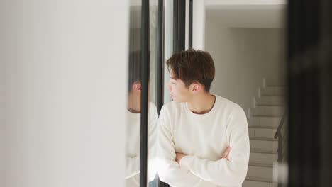 Asian-male-teenager-looking-through-window-with-arms-crossed-in-living-room