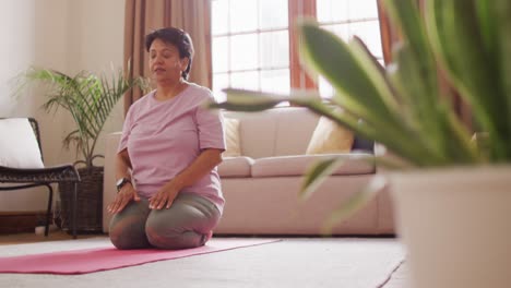 Ältere-Biraziale-Frau,-Die-Yoga-Praktiziert-Und-Im-Wohnzimmer-Meditiert