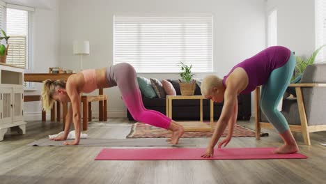 Happy-diverse-female-couple-doing-yoga-together-in-living-room,-stretching