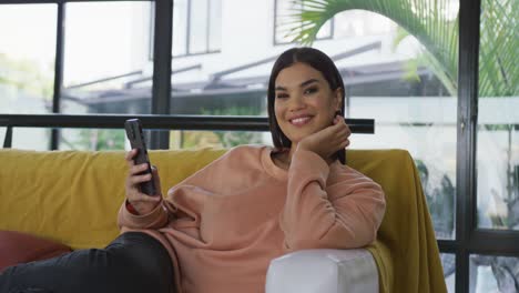 Video-of-happy-biracial-woman-sitting-on-sofa-and-using-smartphone