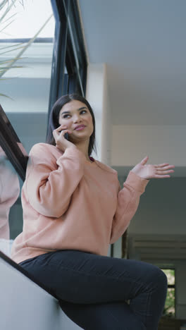 Vertical-video-of-happy-asian-woman-sitting-on-sofa-and-using-smartphone