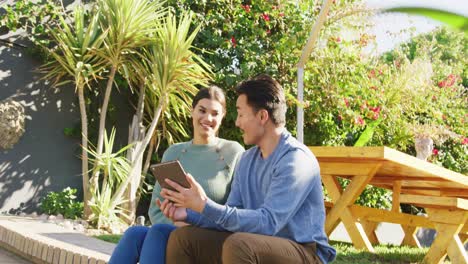 Video-of-happy-diverse-couple-talking-and-using-tablet-in-garden
