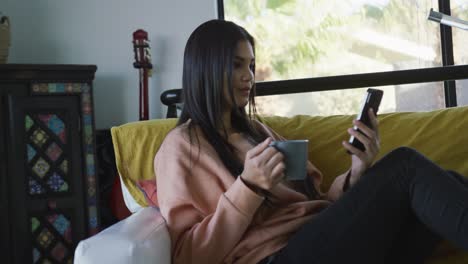 Video-of-biracial-woman-sitting-on-sofa,-drinking-coffee-and-using-smartphone