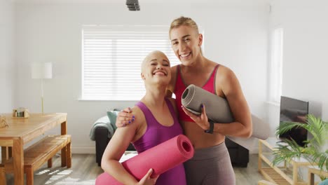 Portrait-of-happy-diverse-female-couple-with-yoga-mats-in-living-room