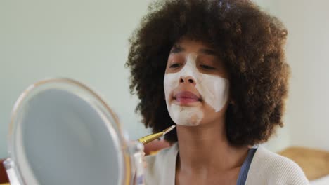 Happy-biracial-woman-applying-beauty-face-mask-with-brush