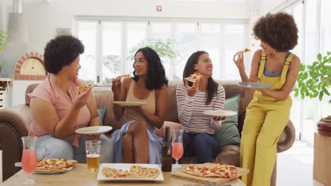 Happy-diverse-female-friends-sitting-on-sofa-in-living-room,-eating-pizza