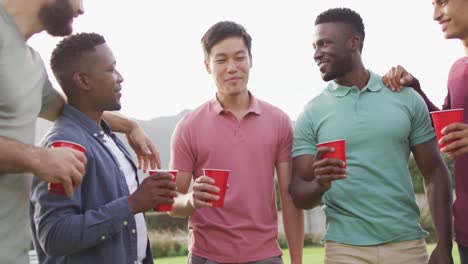 Happy-diverse-male-friends-talking-and-drinking-beer-in-garden-on-sunny-day