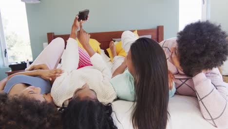 Happy-diverse-female-friends-lying-on-bed-and-using-smartphone-in-bedroom