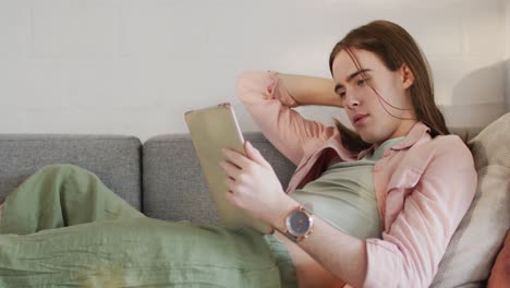 Caucasian-non-binary-transgender-woman-lying-on-sofa,-using-tablet