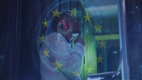 Animation-of-eu-flag-over-african-american-man-using-laptop-in-server-room