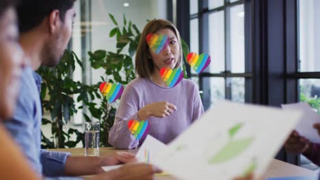 Multiple-rainbow-heart-icons-floating-over-group-of-diverse-office-colleagues-discussing-at-office