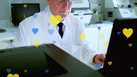 Animation-of-yellow-and-blue-hearts-falling-over-senior-caucasian-male-lab-worker-with-computer