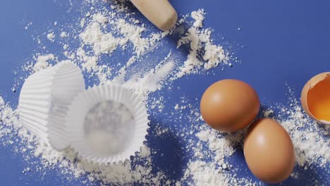 Video-of-baking-ingredients,-muffin-papers,-eggs-and-tools-lying-on-blue-surface-with-flour