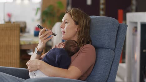 Video-Einer-Kaukasischen-Mutter,-Die-Mit-Ihrem-Neugeborenen-Im-Sessel-Sitzt-Und-Ihr-Smartphone-Benutzt