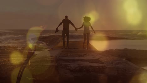 Yellow-spots-of-light-over-african-american-couple-holding-hands-walking-on-the-rocks-near-the-sea