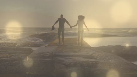 Glowing-spots-of-light-against-african-american-couple-holding-hands-running-on-the-rocks