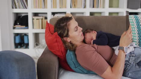 Video-Einer-Kaukasischen-Mutter-Mit-Tablet,-Die-Mit-Ihrem-Neugeborenen-Schlafenden-Baby-Auf-Dem-Sofa-Liegt