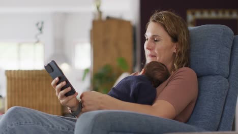 Video-of-caucasian-mother-sitting-in-armchair-with-newborn-baby-and-using-smartphone