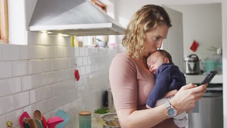 Video-Einer-Kaukasischen-Mutter-Mit-Neugeborenem-Baby-In-Der-Babytrage,-Die-Ihr-Smartphone-In-Der-Küche-Benutzt