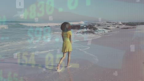 Stock-market-data-processing-against-rear-view-of-african-american-woman-walking-on-the-beach