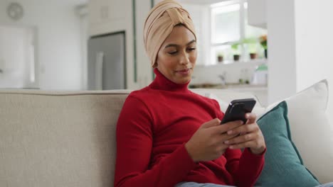 Video-of-happy-biracial-woman-in-hijab-sitting-on-sofa-and-using-smartphone