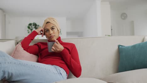 Video-of-biracial-woman-in-hijab-sitting-on-sofa-and-drinking-coffee
