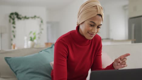 Video-of-happy-biracial-woman-in-hijab-sitting-on-sofa-and-using-laptop